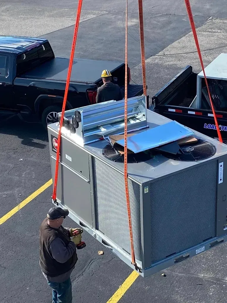 Heavy HVAC unit being lifted by crane, with workers observing nearby.